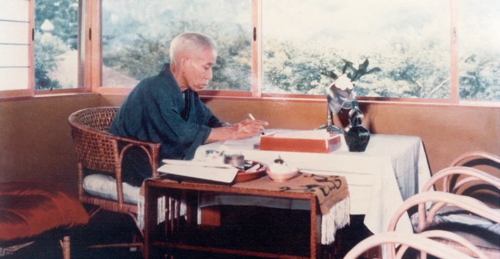 Meishu-Sama sentado &agrave; mesa escrevendo.